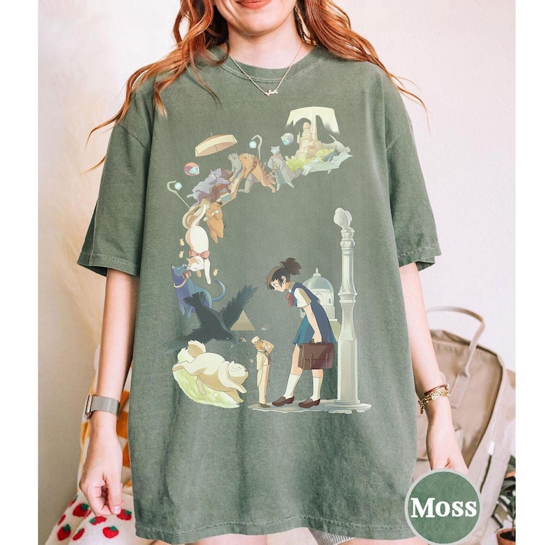 Puede incluir: Camiseta verde musgo con una ilustraci&oacute;n de estilo anime. El gr&aacute;fico representa a una chica con uniforme escolar, rodeada de gatos flotantes, un p&aacute;jaro negro y otros elementos caprichosos. Una farola y un edificio tambi&eacute;n est&aacute;n en el dise&ntilde;o.
