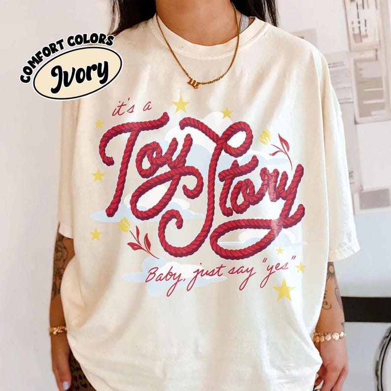 May include: An ivory short-sleeve t-shirt featuring a graphic design with the red rope-textured text "it's a Toy Story" and "Baby, just say "yes"" in cursive. The design includes light blue clouds, yellow stars, and small red floral accents.