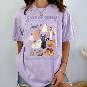 K&ouml;nnte beinhalten: Ein helllila T-Shirt mit einer gerahmten Illustration von Zeichentrickkatzen und -figuren. Der Text "CATS OF GHIBLI" ist &uuml;ber der Illustration aufgedruckt. Das Design zeigt verschiedene Katzenfiguren in verschiedenen Posen und Farben.