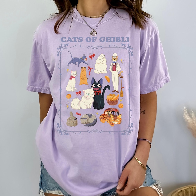 K&ouml;nnte beinhalten: Ein helllila T-Shirt mit einer gerahmten Illustration von Zeichentrickkatzen und -figuren. Der Text "CATS OF GHIBLI" ist &uuml;ber der Illustration aufgedruckt. Das Design zeigt verschiedene Katzenfiguren in verschiedenen Posen und Farben.