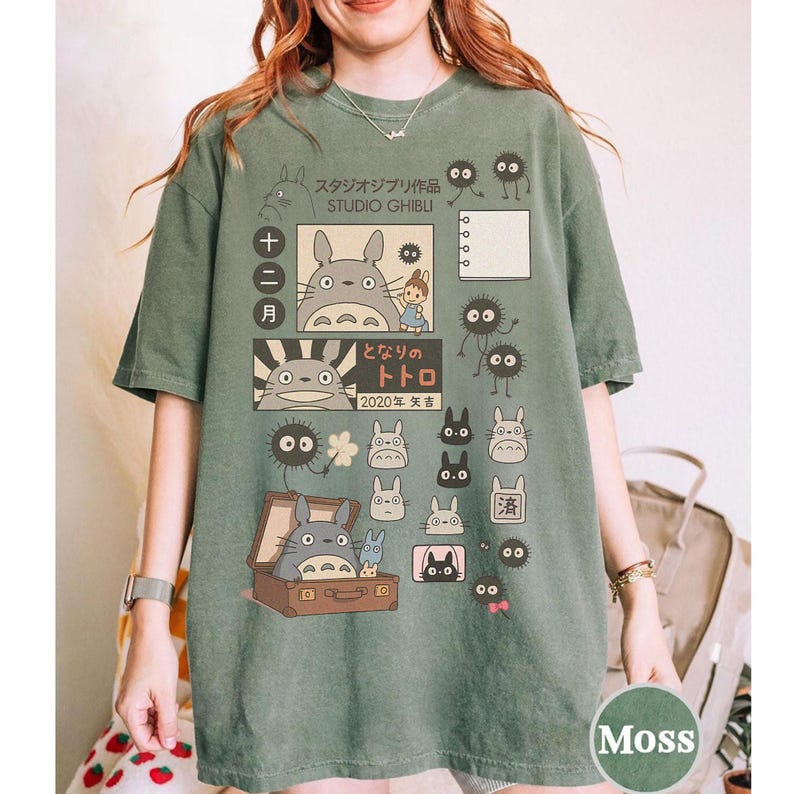 May include: A moss green t-shirt featuring a Studio Ghibli design with characters from the movie "My Neighbor Totoro." The design includes Japanese text, Totoro, soot sprites, and other characters. The shirt has a classic crew neck and short sleeves.