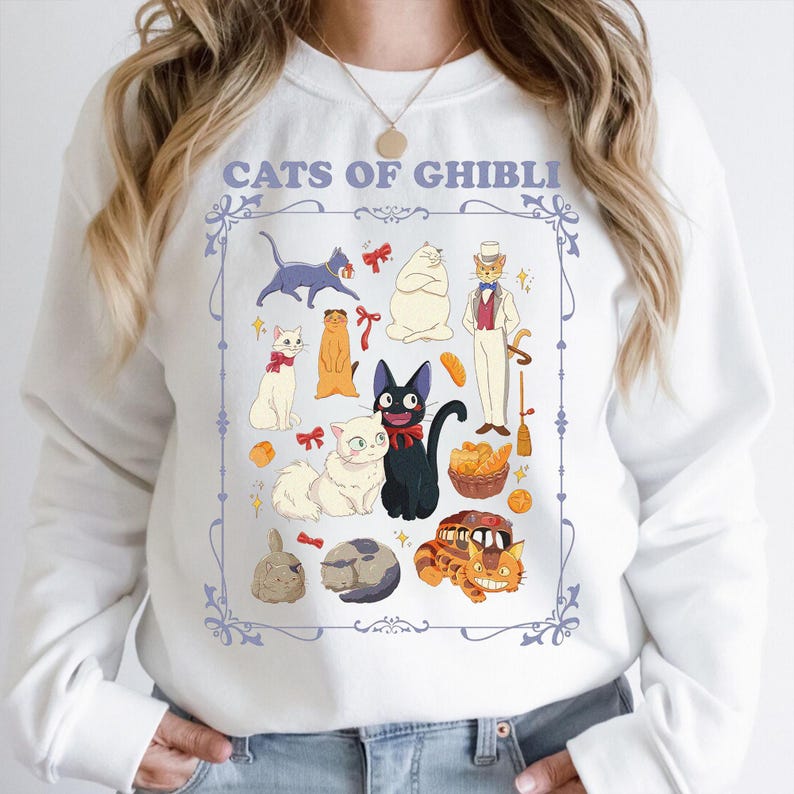 K&ouml;nnte beinhalten: Wei&szlig;es Sweatshirt mit einer farbenfrohen Illustration von Katzenfiguren aus Studio Ghibli-Filmen. Der Text "CATS OF GHIBLI" ist oben aufgedruckt. Das Design umfasst verschiedene Katzenfiguren, Schleifen und andere dekorative Elemente.
