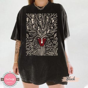 以下が含まれることがあります： 黒いTシャツ。赤い顔と白と黒のディテールを持つ森の精霊の詳細なグラフィックが特徴です。デザインには、小さなフィギュアのある木のような構造が含まれています。「Black」の文字がTシャツにプリントされています。