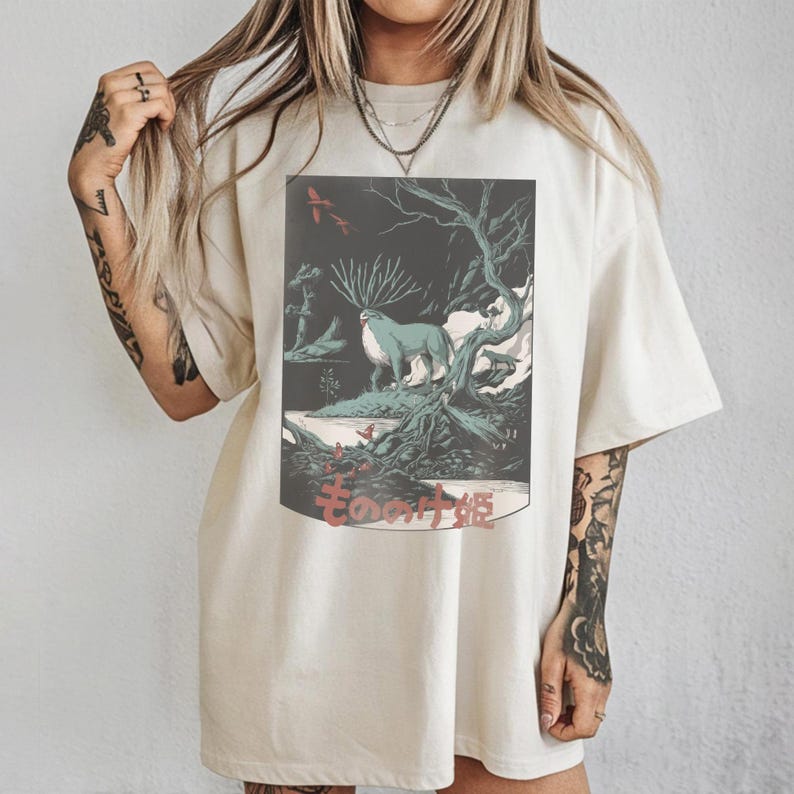 May include: Cream-colored t-shirt featuring a graphic print of a forest scene with a blue animal and Japanese text. The artwork includes trees, birds, and a dark background. The shirt has short sleeves and a relaxed fit.