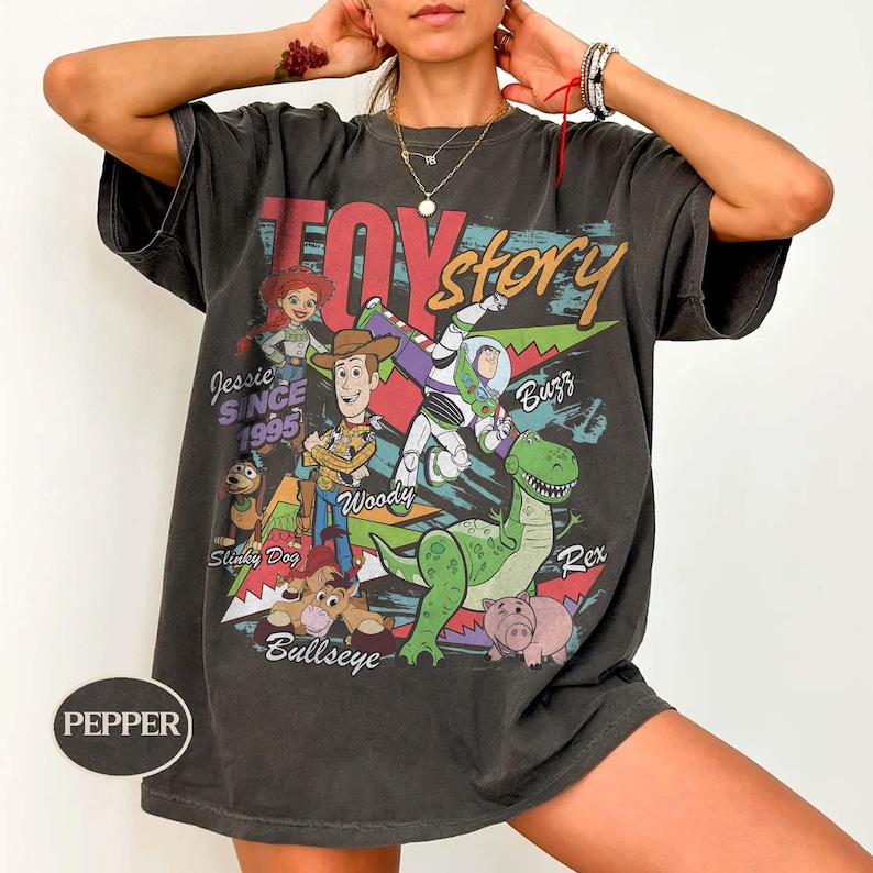 May include: A charcoal gray short-sleeve t-shirt featuring a graphic print of Toy Story characters including Woody, Buzz Lightyear, Jessie, Rex, Slinky Dog, and Bullseye. The text reads Toy Story, Jessie Since 1995, Woody, Buzz, Rex, and Bullseye.