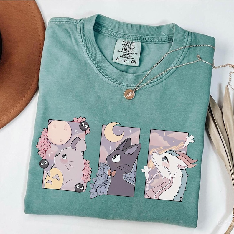 May include: A seafoam green t-shirt featuring three framed anime-style illustrations. The illustrations depict a gray creature with a yellow belly, a black cat with a bow tie, and a white dragon. A gold necklace and a brown hat are also visible.