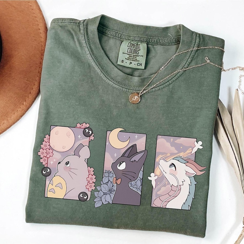 May include: Olive green t-shirt with a graphic featuring three anime-style characters in rectangular frames. The shirt has a crew neck and short sleeves. A gold necklace with a pendant rests on the shirt. The shirt is labeled "Comfort Colors".