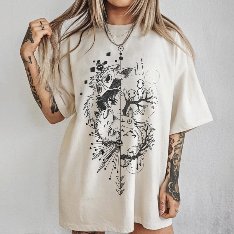 Puede incluir: Camiseta color crema con un dise&ntilde;o gr&aacute;fico en blanco y negro. El dise&ntilde;o est&aacute; dividido verticalmente, mostrando un personaje con una m&aacute;scara en un lado y una escena de bosque con figuras en el otro. Formas geom&eacute;tricas y flechas completan el dise&ntilde;o.