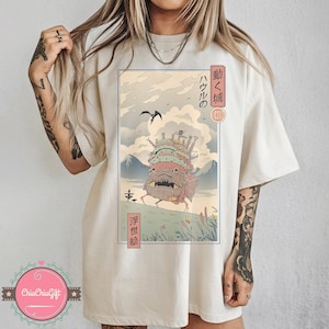 Pu&ograve; includere: T-shirt color crema con un'illustrazione incorniciata del castello in movimento del film d'animazione "Il castello errante di Howl". L'opera d'arte include testo giapponese e un paesaggio con montagne, un uccello e il castello. La maglietta &egrave; di vestibilit&agrave; ampia.