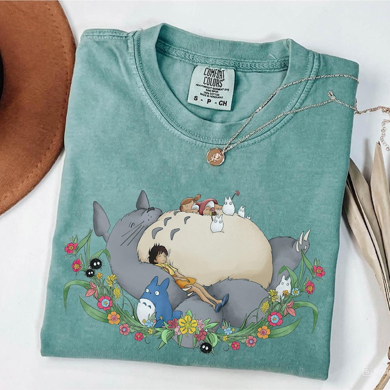 Puede incluir: Una camiseta Comfort Colors verde azulado con un gr&aacute;fico de Totoro y otros personajes de la pel&iacute;cula de animaci&oacute;n Mi Vecino Totoro. El dise&ntilde;o incluye una corona floral y la etiqueta Comfort Colors.