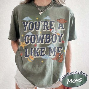 May include: A moss green short-sleeve t-shirt featuring the text YOU'RE A COWBOY LIKE ME in dark blue letters. The graphic includes a brown cowboy hat, a rocket ship, a yellow sheriff star, a colorful ball, and small stars and clouds.