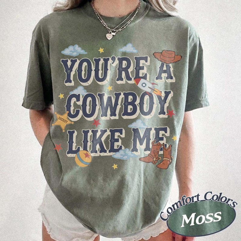 May include: A moss green short-sleeve t-shirt featuring the text YOU'RE A COWBOY LIKE ME in dark blue letters. The graphic includes a brown cowboy hat, a rocket ship, a yellow sheriff star, a colorful ball, and small stars and clouds.