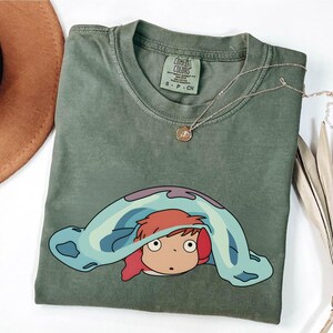 May include: Olive green Comfort Colors t-shirt featuring a cartoon character design. The graphic shows a character with red hair peeking out from under a blue and purple blanket. A gold necklace is draped over the shirt.