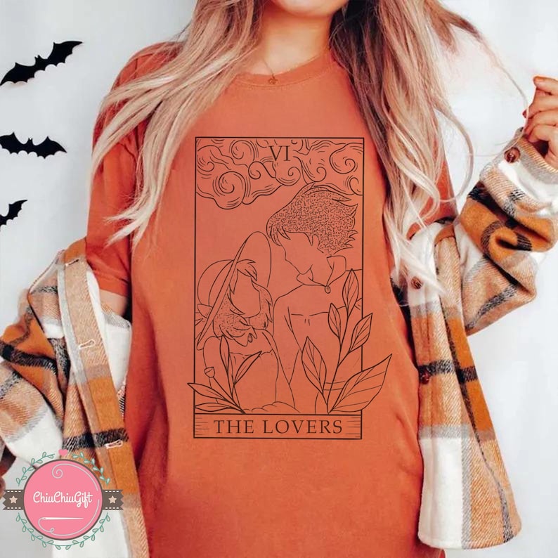 Puede incluir: Camiseta naranja con un dibujo lineal negro de una carta del tarot, "Los Amantes". El dise&ntilde;o incluye dos figuras, hojas y el n&uacute;mero romano VI. Se ven recortes de murci&eacute;lagos negros en la esquina superior izquierda.