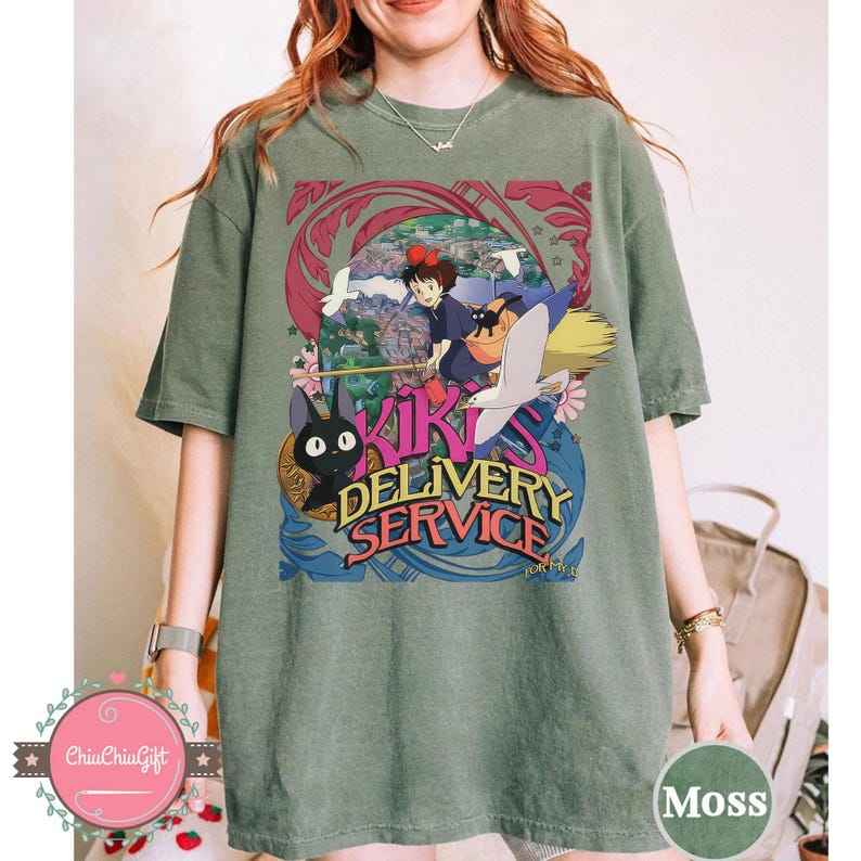 May include: Moss green t-shirt with a vibrant graphic from "Kiki's Delivery Service." The design showcases the text "Kiki's Delivery Service" alongside illustrations of Kiki, Jiji the cat, and a broom. The t-shirt has a relaxed fit.