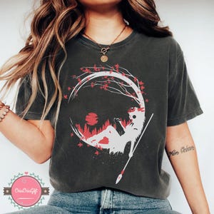 May include: Dark gray t-shirt featuring a white and red graphic of a figure reclining under a tree with a spear. The design includes a red sun and forest scene within a circular frame. The shirt has a crew neck.