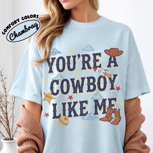 K&ouml;nnte beinhalten: Ein hellblaues T-Shirt mit dem dunkelblauen Text "YOU'RE A COWBOY LIKE ME", umgeben von Illustrationen eines braunen Cowboyhuts, einer Rakete, eines gelben Sheriffsterns, eines Balls sowie kleinen roten Sternen und hellblauen Wolken. Ein Logo zeigt "COMFORT COLORS Chambray".