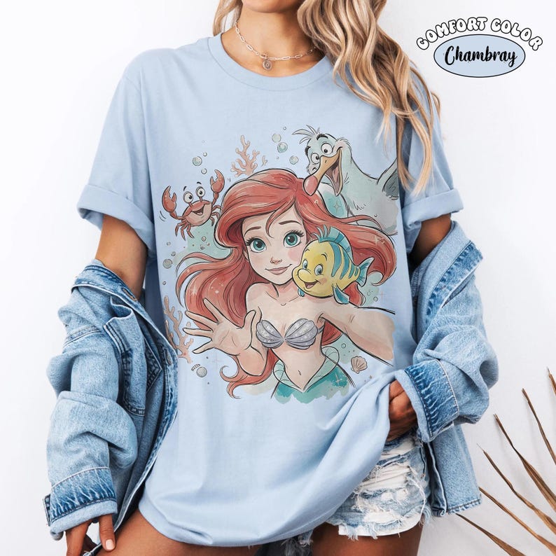 Puede incluir: Una camiseta de manga corta azul claro con un estampado gr&aacute;fico de una sirena de pelo rojo, un pez amarillo y azul, un peque&ntilde;o cangrejo rojo y una gaviota blanca. El texto COMFORT COLOR Chambray est&aacute; impreso en la esquina superior derecha.