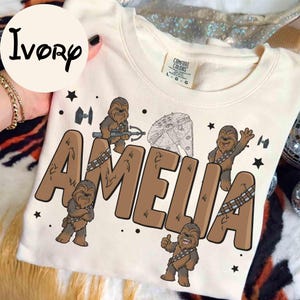 Può includere: Felpa color avorio con il nome "AMELIA" in lettere marroni, illustrazioni di Chewbacca e grafica a tema Star Wars. La maglia ha un'etichetta Comfort Colors.