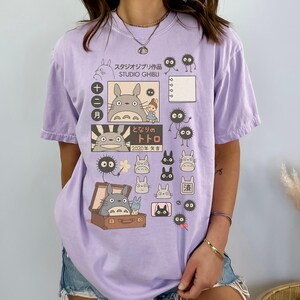 May include: A light purple t-shirt featuring Studio Ghibli characters and Japanese text. The design includes illustrations of Totoro, soot sprites, and other characters from the animated film. The shirt has a relaxed fit and short sleeves.