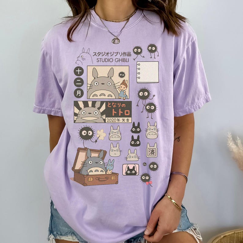 May include: A light purple t-shirt featuring Studio Ghibli characters and Japanese text. The design includes illustrations of Totoro, soot sprites, and other characters from the animated film. The shirt has a relaxed fit and short sleeves.