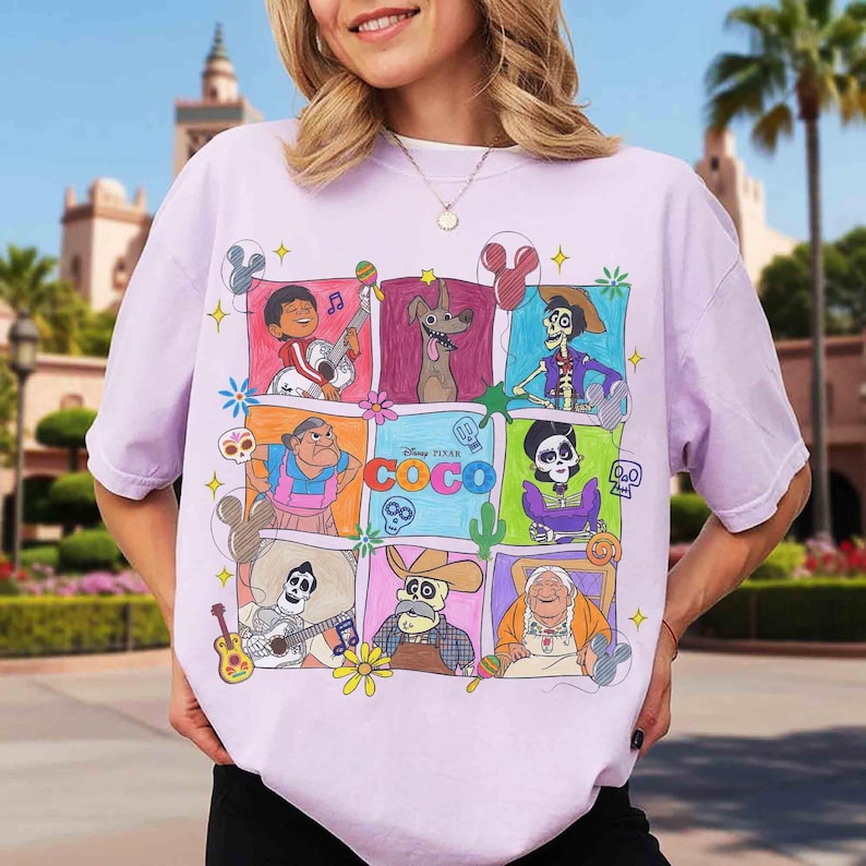 May include: A light purple t-shirt featuring a colorful graphic of characters from the Disney Pixar movie "Coco." The design includes various characters in square frames, with the movie's logo prominently displayed. The shirt has short sleeves.