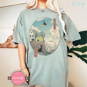 Op de afbeelding: Saliegroen T-shirt met een cirkelvormige afbeelding in Japanse kunststijl. De afbeelding toont een heks op een bezem, een zwarte kat, gebouwen en Japanse tekst. Het shirt heeft korte mouwen en een losse pasvorm.