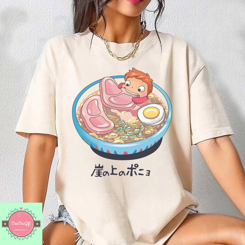 May include: Beige t-shirt featuring a cartoon illustration of a ramen bowl with a character from the animated film Ponyo. The bowl is blue with a detailed design, and the text "崖の上のポニョ" is below the bowl.