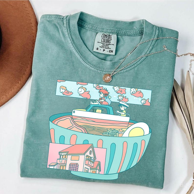 Puede incluir: Una camiseta Comfort Colors verde azulado con un gr&aacute;fico colorido de un cuenco de ramen, un coche y personajes. El dise&ntilde;o incluye una casa, fideos y un huevo. Un collar dorado est&aacute; drapeado sobre la camisa.