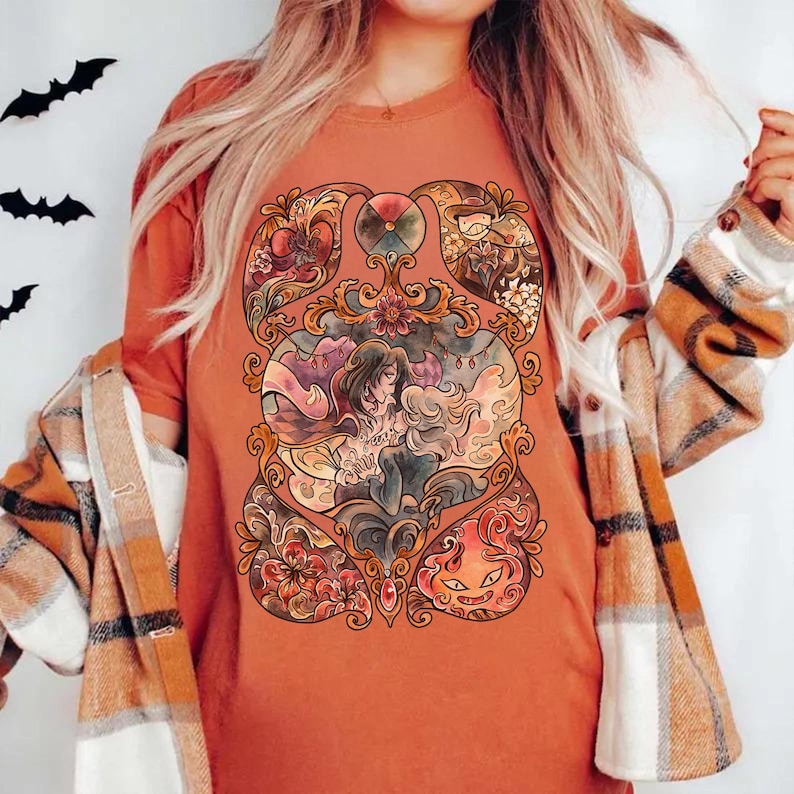 May include: Burnt orange t-shirt featuring a detailed, colorful illustration. The artwork depicts a central figure with flowing hair, surrounded by floral and fantasy elements. The design has a stained-glass window aesthetic, with a Halloween theme.