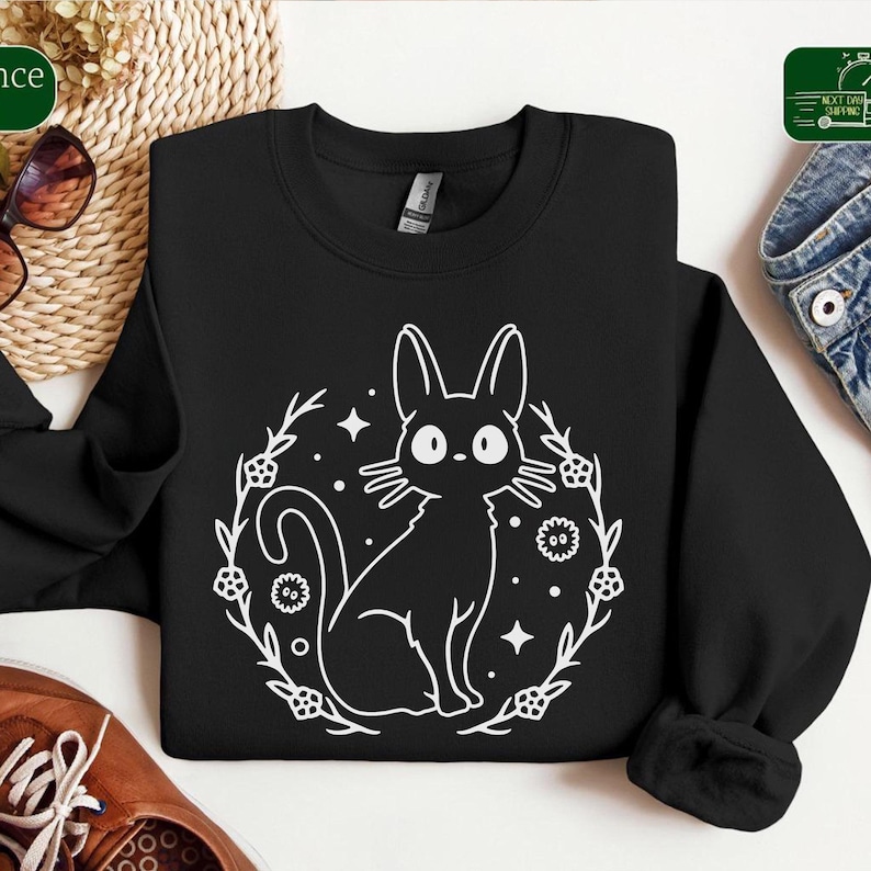 May include: Black sweatshirt featuring a white graphic of a cat inside a floral wreath with stars and dots. The cat has large eyes and long ears. The sweatshirt is made of a soft material.