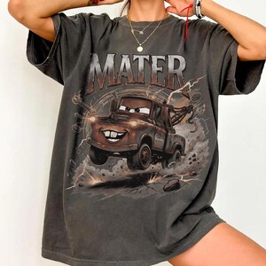 Pode incluir: Uma t-shirt cinzento-carv&atilde;o de manga curta com uma impress&atilde;o gr&aacute;fica de um reboque castanho com o texto MATER arqueado por cima. O design inclui rel&acirc;mpagos estilizados, nuvens de poeira e detritos em torno do ve&iacute;culo.