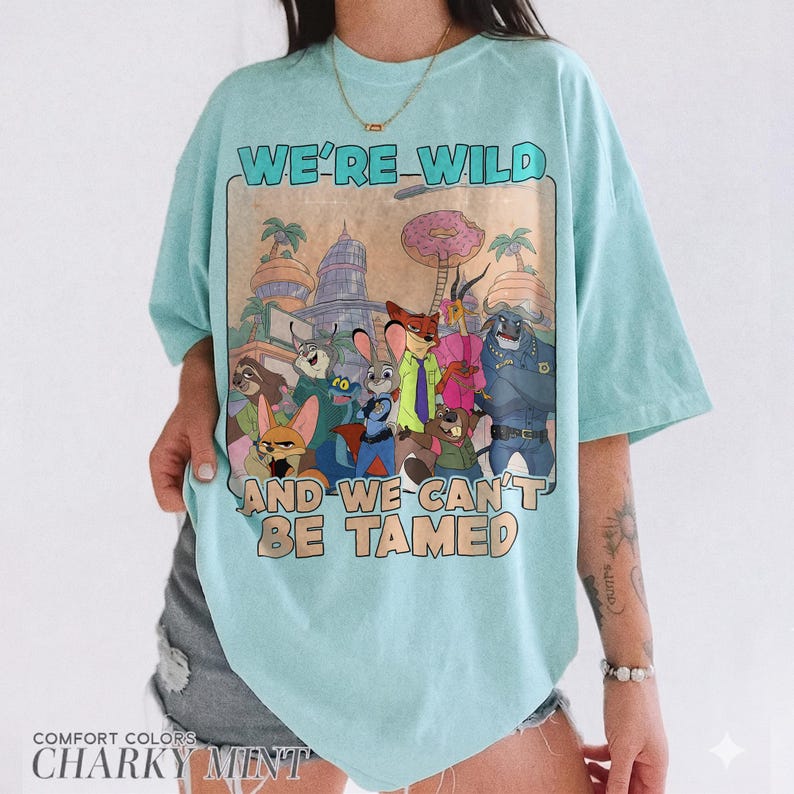 May include: A light mint green short-sleeve t-shirt featuring a graphic print of various animated animal characters from Zootopia. The text WE'RE WILD AND WE CAN'T BE TAMED is printed in a bold, light-colored font above and below the central illustration.