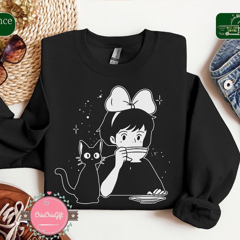 Pu&ograve; includere: Felpa nera con una grafica bianca di una ragazza con un fiocco che beve il t&egrave;, accompagnata da un gatto nero. Il design include stelle e croci. Un capo casual e confortevole.