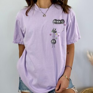 May include: A light purple t-shirt with a pocket featuring a cartoon design of three black, fluffy creatures with eyes and legs. The creatures are stacked, with star accents. The shirt is short-sleeved and has a relaxed fit.