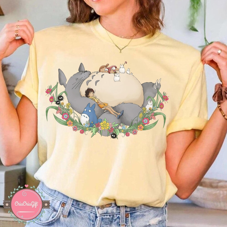 Puede incluir: Camiseta amarilla clara con un gr&aacute;fico de un gran personaje gris de Totoro, personajes m&aacute;s peque&ntilde;os y una ni&ntilde;a acostada sobre su vientre, rodeados de flores coloridas. El dise&ntilde;o est&aacute; centrado en la camiseta.