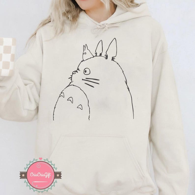 Puede incluir: Una sudadera con capucha color crema con un dibujo lineal negro de un personaje de Totoro de la pel&iacute;cula de animaci&oacute;n Mi vecino Totoro. El dise&ntilde;o est&aacute; en la parte delantera. La sudadera tiene un bolsillo de canguro y una capucha con cord&oacute;n.