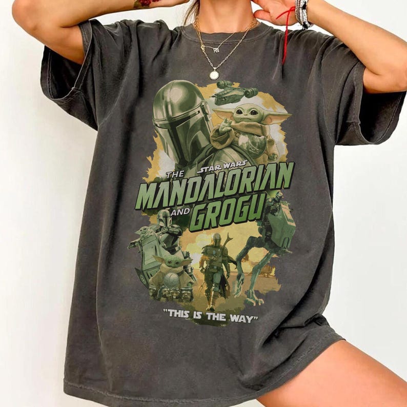 May include: A charcoal gray short-sleeve t-shirt featuring a graphic print of The Mandalorian and Grogu. The design includes the text "THE MANDALORIAN AND GROGU" and "THIS IS THE WAY" in green lettering, surrounded by various character illustrations.
