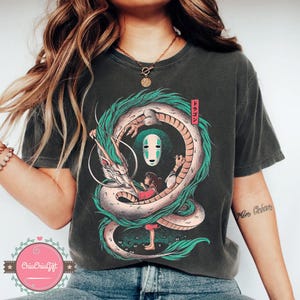 May include: Dark gray t-shirt featuring a colorful illustration of a dragon encircling a girl and a masked figure. The dragon is white and green, with the girl in a red dress. Japanese text is in a red rectangle.