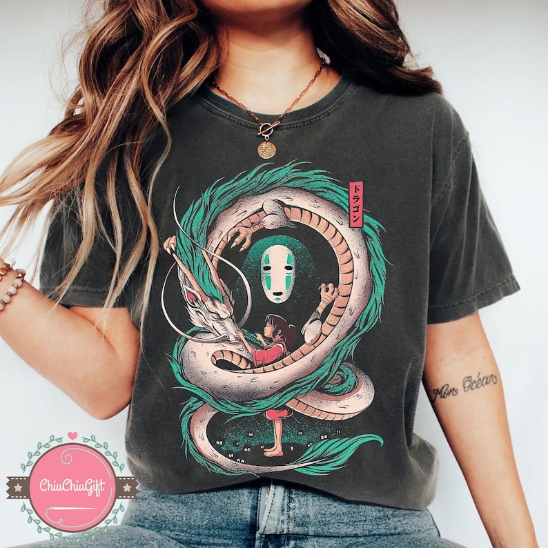 May include: Dark gray t-shirt featuring a colorful illustration of a dragon encircling a girl and a masked figure. The dragon is white and green, with the girl in a red dress. Japanese text is in a red rectangle.