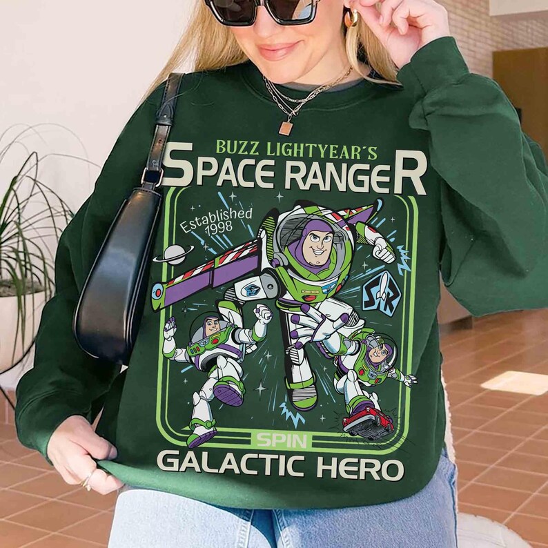 May include: Dark green sweatshirt with a Buzz Lightyear graphic. The design includes the text "Buzz Lightyear's Space Ranger" and "Galactic Hero". The graphic is in shades of green, purple, white, and red. The sweatshirt is a crew neck style.