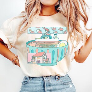 Puede incluir: Camiseta color crema con una ilustraci&oacute;n caprichosa. El dise&ntilde;o representa un gran cuenco de ramen con un barco y dos figuras. Arriba, nadan peces de dibujos animados. Abajo, se muestra una casa. La camiseta es de material suave.