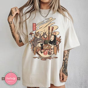 May include: Cream-colored t-shirt featuring a colorful graphic of a ramen bowl with animated characters. The design includes a bowl of noodles, chopsticks, and the Japanese text "ラーメン".