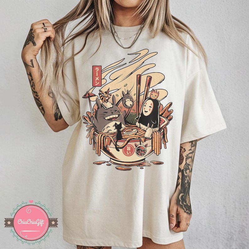 May include: Cream-colored t-shirt featuring a colorful graphic of a ramen bowl with animated characters. The design includes a bowl of noodles, chopsticks, and the Japanese text "ラーメン".