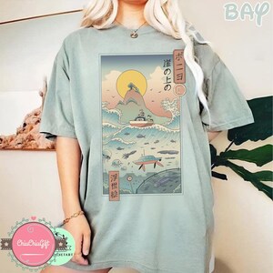 May include: Sage green t-shirt featuring a Japanese-inspired graphic. The graphic depicts a boat on ocean waves, a submarine, and a rising sun. Japanese text is also included in the design.