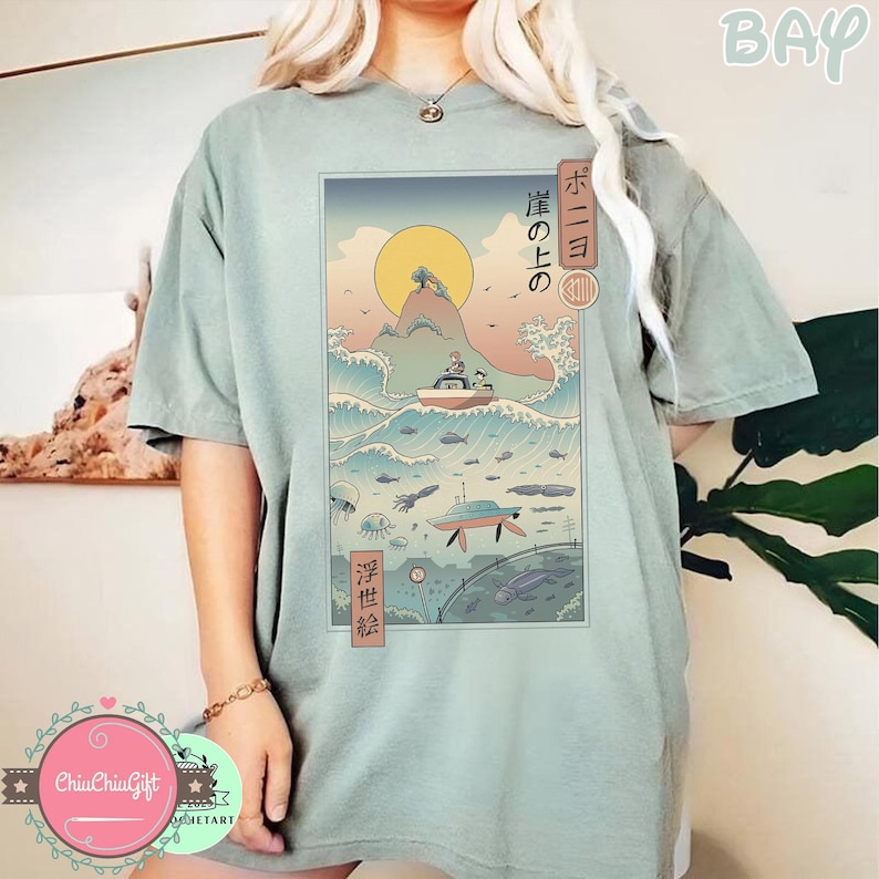 May include: Sage green t-shirt featuring a Japanese-inspired graphic. The graphic depicts a boat on ocean waves, a submarine, and a rising sun. Japanese text is also included in the design.