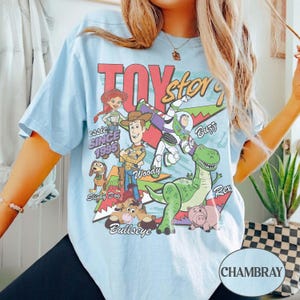 May include: A light blue t-shirt featuring a graphic print of Toy Story characters including Woody, Buzz Lightyear, Jessie, Rex, Slinky Dog, and Bullseye. The design includes the text Toy Story, Since 1995, and the names of the characters in various fonts.