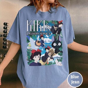 May include: Blue jean-colored t-shirt featuring the text "Kiki's Delivery Service" above a colorful graphic of characters from the animated film. The design includes a witch on a broom, a black cat, and other animated figures.