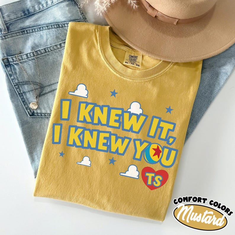 May include: A mustard yellow short-sleeve t-shirt featuring the blue text I KNEW IT, I KNEW YOU with a red heart containing the letters TS and a colourful ball graphic. The shirt includes white cloud and blue star accents, with a Comfort Colors label.
