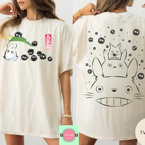May include: Ivory-colored t-shirt featuring a front graphic of a white cartoon character with a green leaf and black soot sprites, and Japanese text. The back of the shirt has a large graphic of the same character with two smaller characters and more soot sprites.